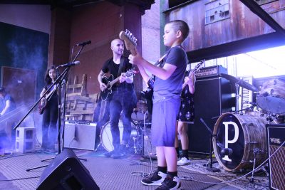 Group of 3 child musicians perform with an adult guitarist surrounded by smoke and cool lighting.
